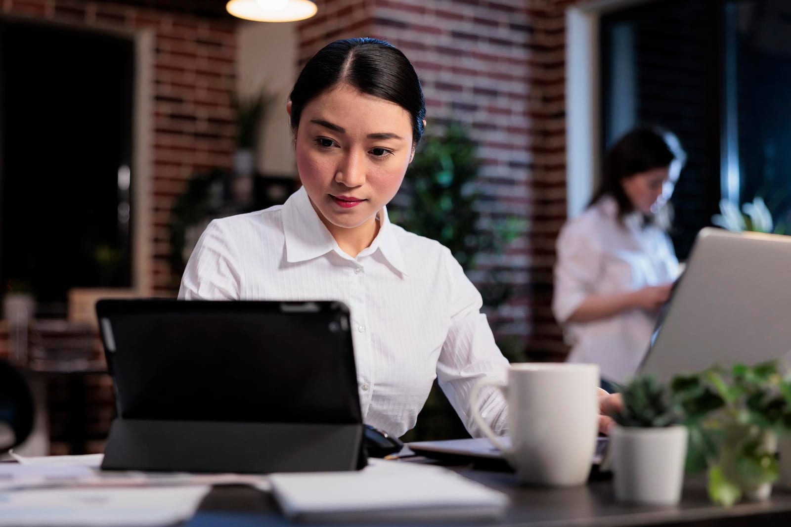 デスクでタブレットを確認する女性と、背後で作業する同僚。